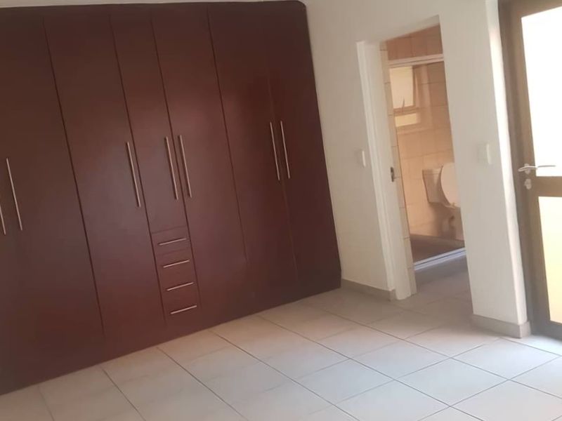 A clean, empty bedroom featuring tiled flooring and dark wood built-in cupboards, with an adjoining en-suite bathroom visible through the doorway.