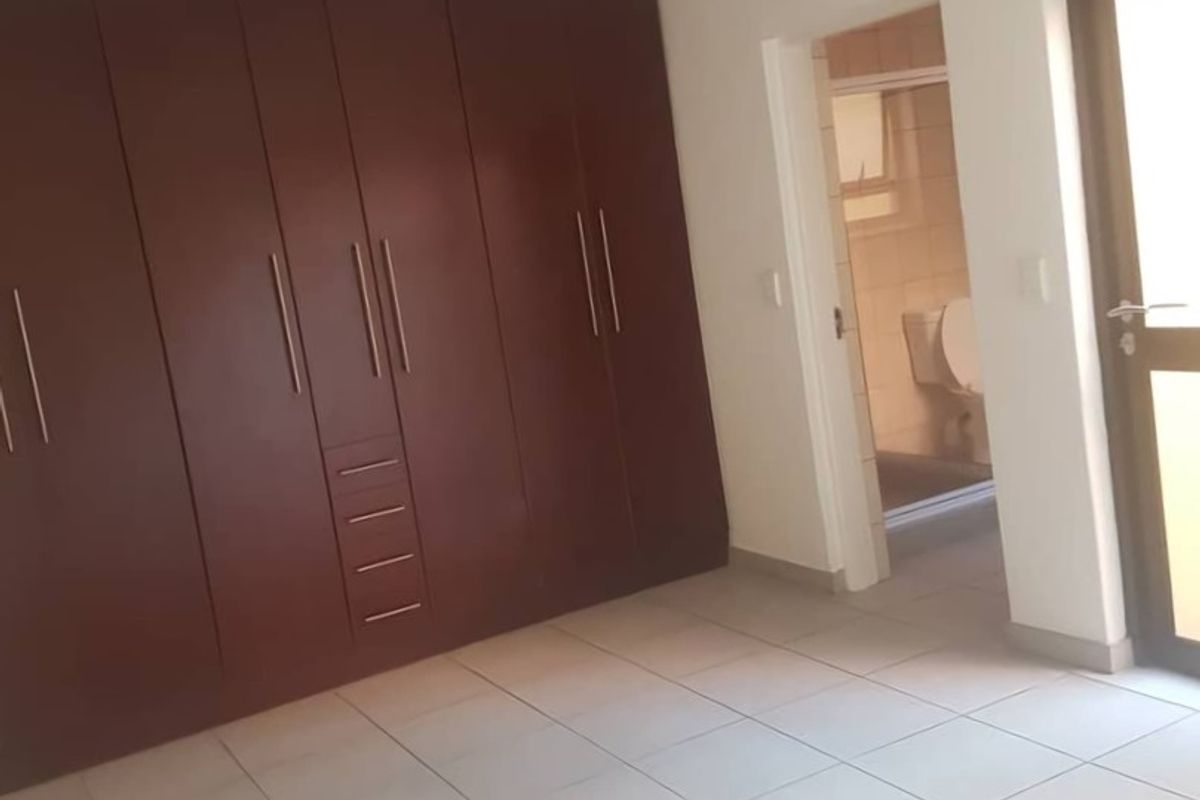 A clean, empty bedroom featuring tiled flooring and dark wood built-in cupboards, with an adjoining en-suite bathroom visible through the doorway.