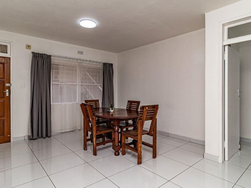 A bright dining area featuring large white floor tiles, a wooden entrance door, and a window with security bars.
