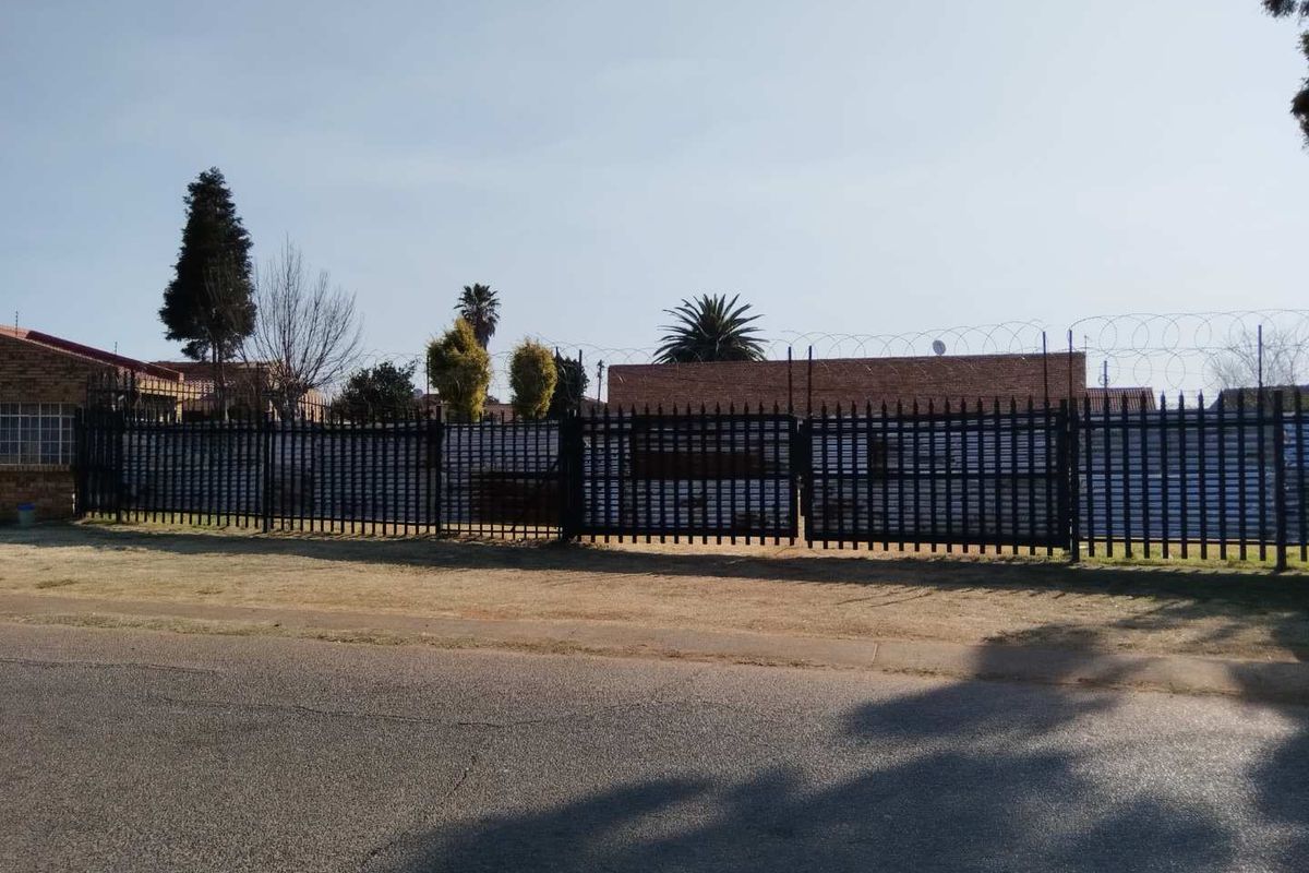 A street-level view of a residential property featuring secure metal perimeter fencing topped with razor wire and a gated entrance.