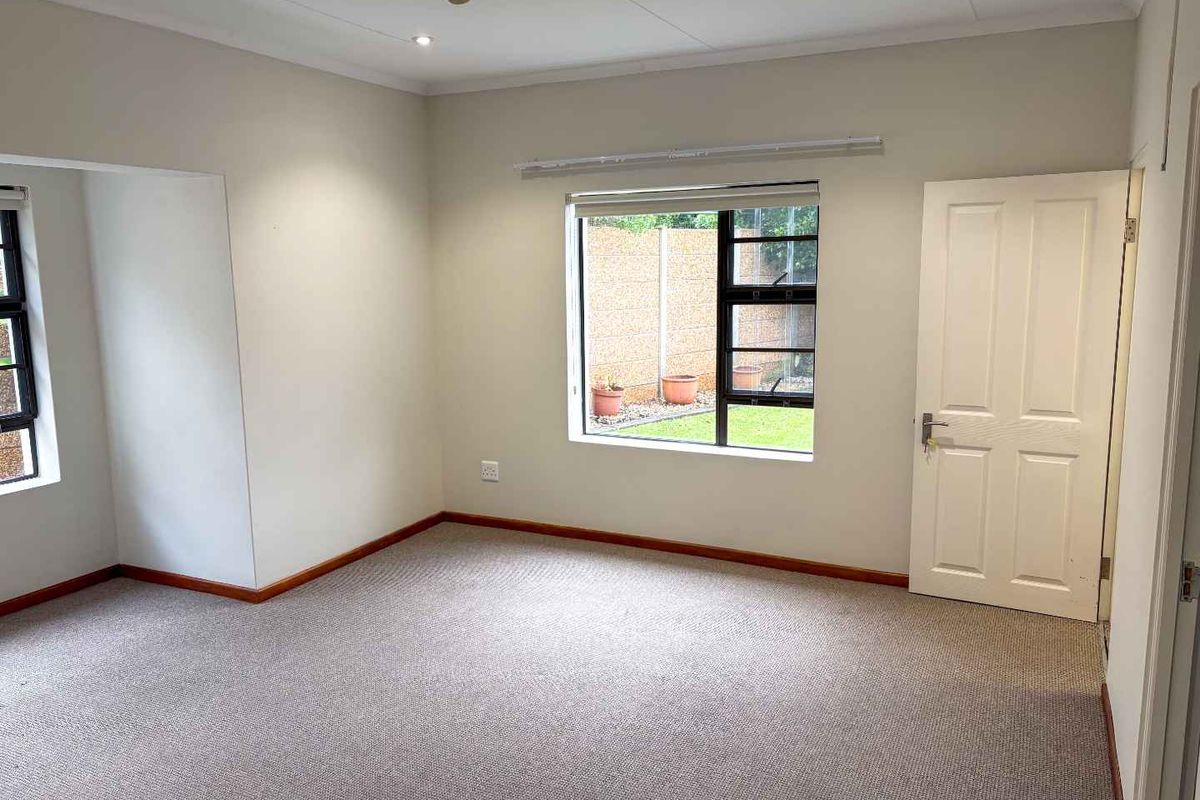 A bright, empty bedroom featuring neutral walls, carpeted flooring, a ceiling fan, and large windows providing natural light and garden views.