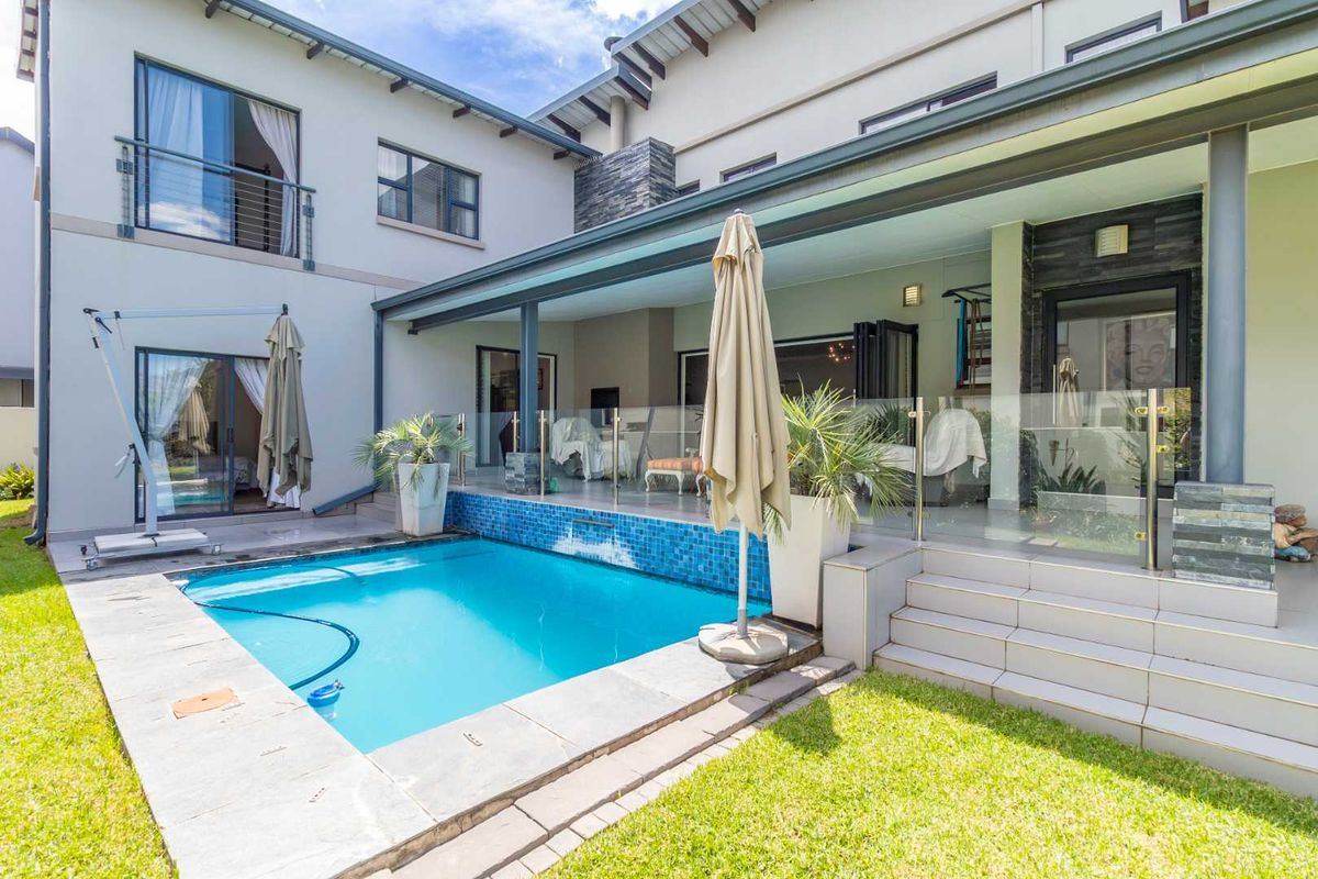 A well-maintained rear exterior featuring a covered patio, glass balustrades, and a swimming pool with mosaic tile detailing.