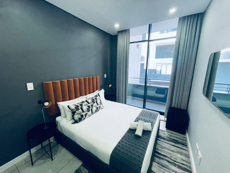 Modern bedroom featuring dark accent wall, floor-to-ceiling windows with balcony access, and recessed lighting, creating a contemporary and comfortable living space.