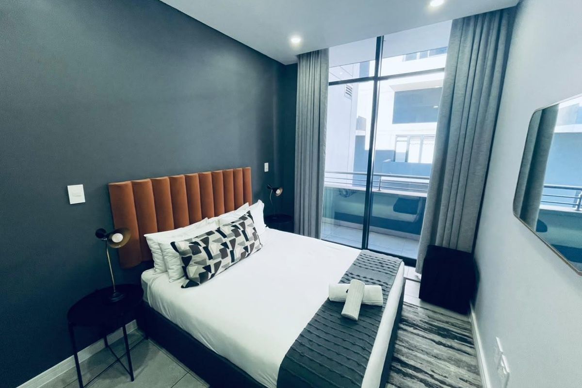 Modern bedroom featuring dark accent wall, floor-to-ceiling windows with balcony access, and recessed lighting, creating a contemporary and comfortable living space.
