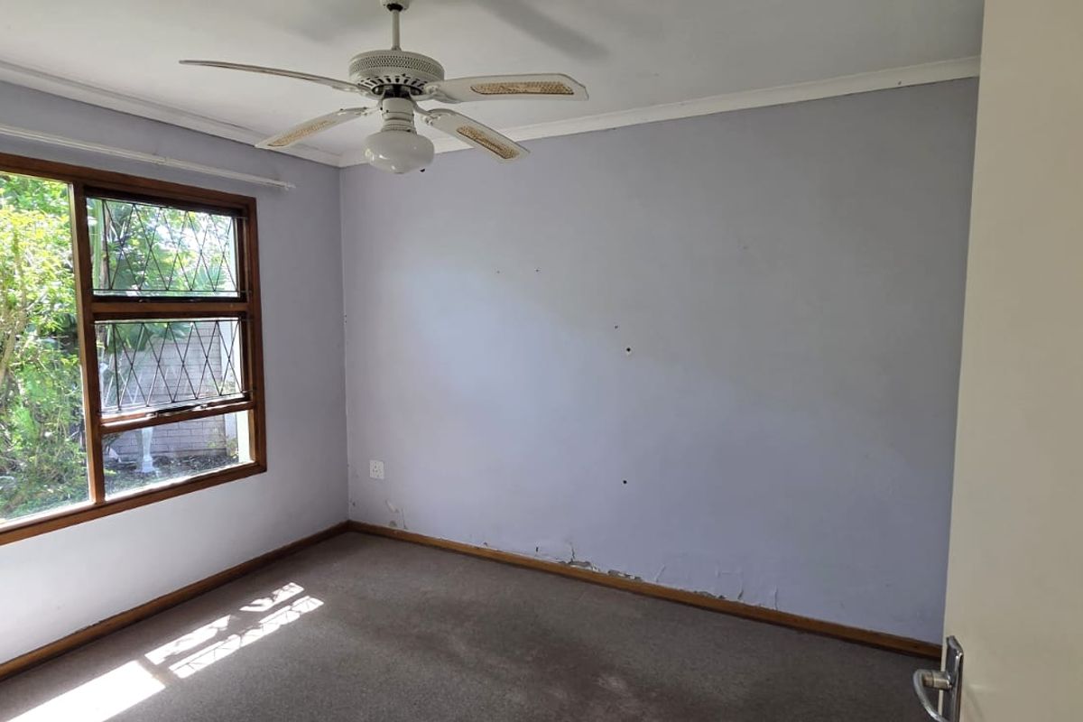 An empty bedroom featuring a ceiling fan, carpeted flooring, and a large leaded glass window with visible wall surface damage.