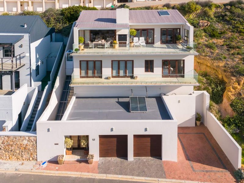 Aerial view of a multi-level modern residence featuring a double garage, paved driveway, expansive balconies with glass balustrades, and solar water heating panels.