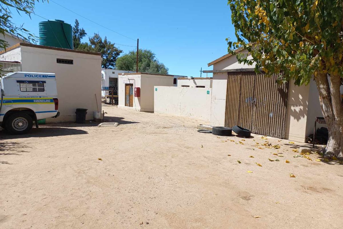 Exterior view of a property featuring multiple outbuildings, a metal garage door, and a large water tank on a sandy, unpaved yard.