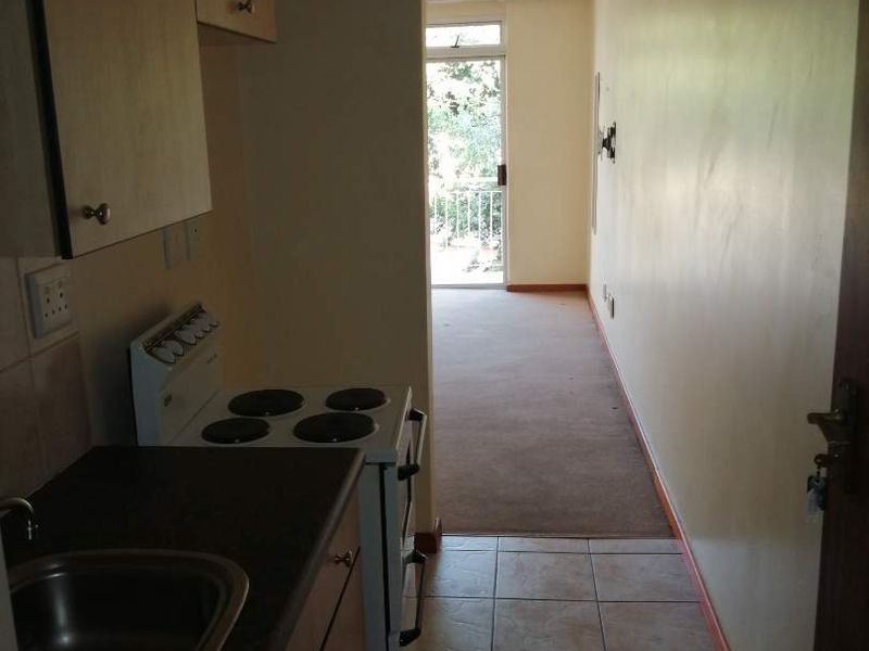 A compact galley-style kitchen featuring built-in cabinetry and a freestanding stove, leading into an adjacent carpeted living area with access to a balcony.