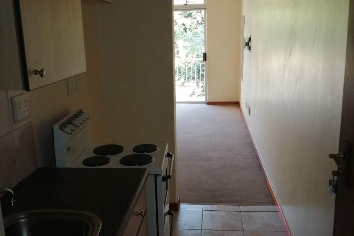 A compact galley-style kitchen featuring built-in cabinetry and a freestanding stove, leading into an adjacent carpeted living area with access to a balcony.