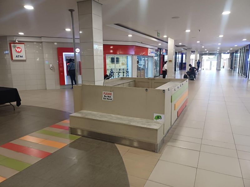 A view of a retail shopping centre hallway featuring a central kiosk, tiled flooring, recessed lighting, and an integrated ATM facility.