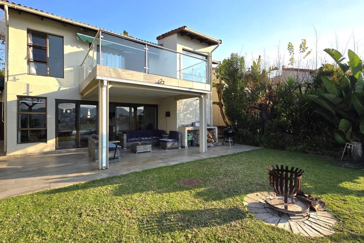 Rear view of the townhouse featuring a covered tiled patio, an upper-level balcony with glass balustrades, a built-in braai, and a landscaped garden with a fire pit.