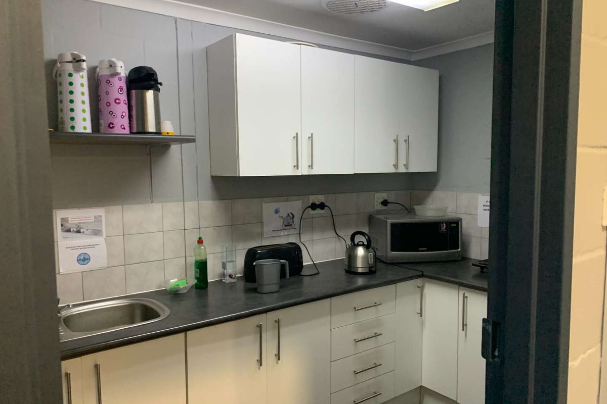 A functional office kitchenette featuring white cabinetry, dark laminate countertops, a stainless steel sink, and tiled splashbacks.