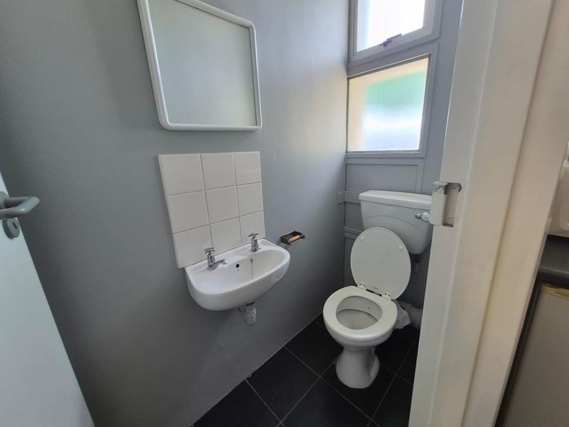 A compact office restroom featuring a wall-mounted basin, toilet, tiled splashback, and dark floor tiles with natural light from casement windows.