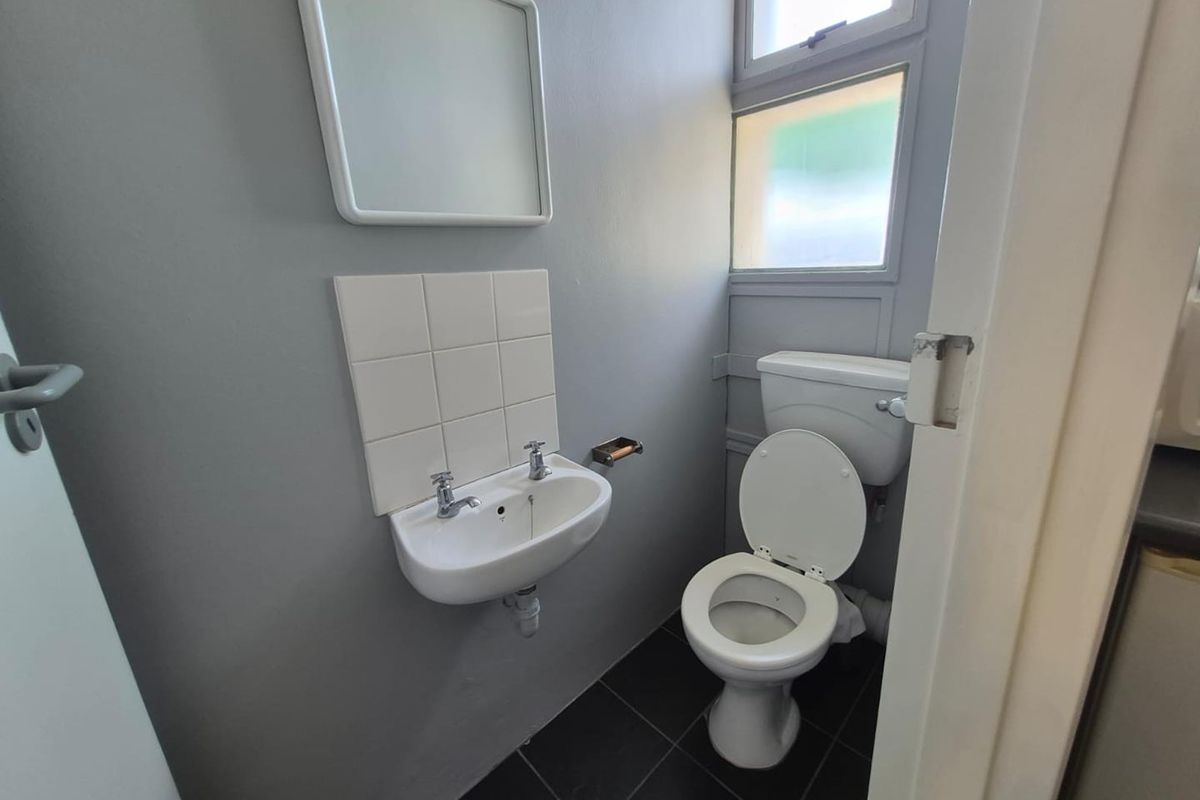 A compact office restroom featuring a wall-mounted basin, toilet, tiled splashback, and dark floor tiles with natural light from casement windows.