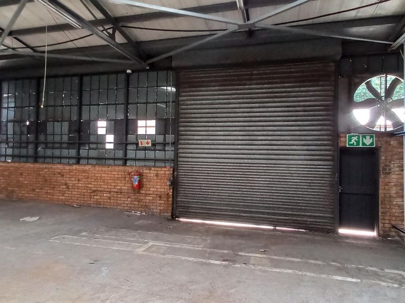Spacious industrial utility area featuring high ceilings with exposed steel trusses, corrugated metal walls, a large roller shutter door, and an industrial extractor fan.