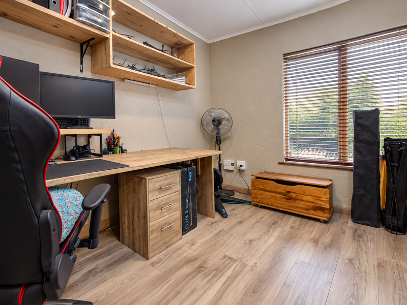A functional home office featuring laminate flooring, built-in wooden shelving, a custom desk, and large windows with venetian blinds.