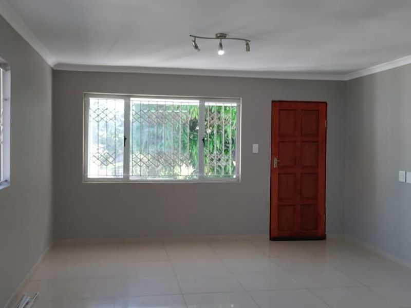A spacious, light-filled living area featuring neutral grey walls, large windows with security gates, tiled flooring, and a classic timber entrance door.