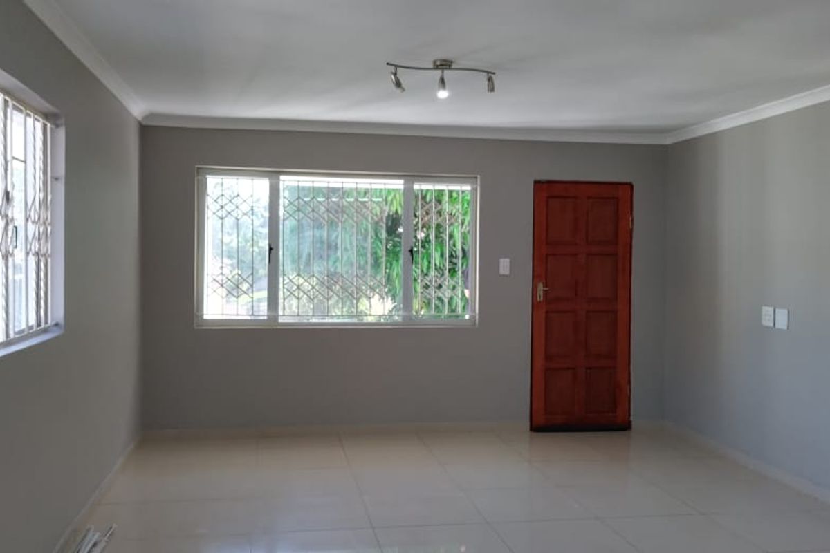 A spacious, light-filled living area featuring neutral grey walls, large windows with security gates, tiled flooring, and a classic timber entrance door.
