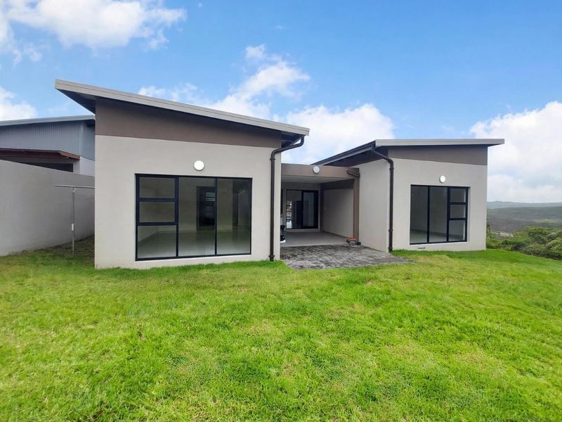 Rear view of a modern single-story house featuring a paved patio, large aluminium-framed windows, and a contemporary skillion roof design set against a grassy lawn.