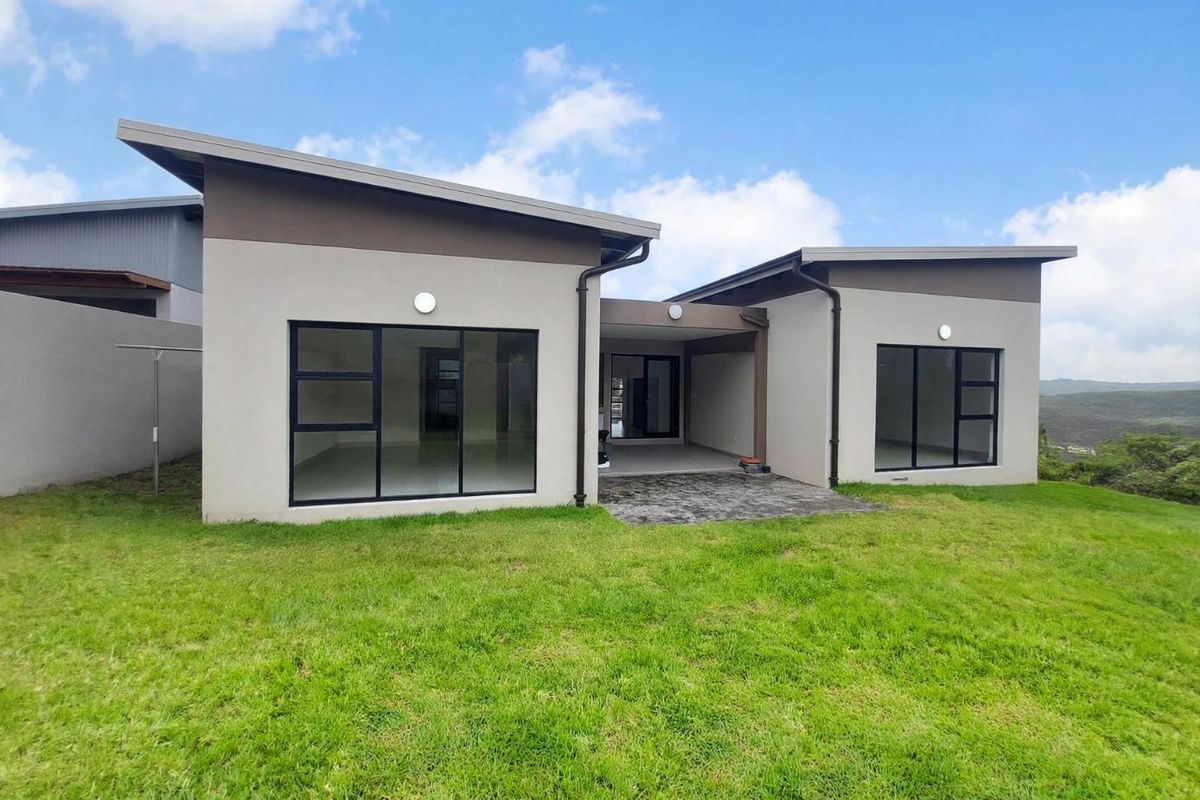 Rear view of a modern single-story house featuring a paved patio, large aluminium-framed windows, and a contemporary skillion roof design set against a grassy lawn.