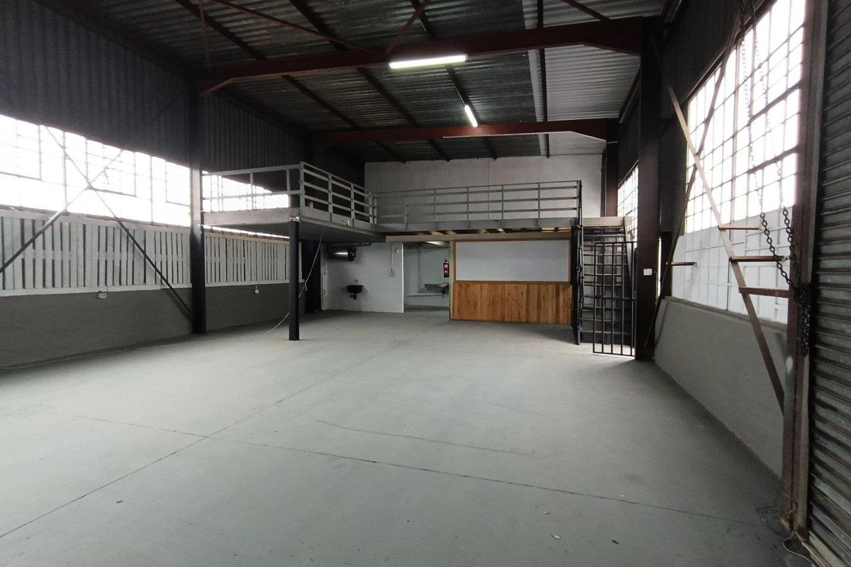 Spacious industrial utility area featuring a mezzanine level, exposed steel roof trusses, concrete flooring, and large industrial windows providing ample natural light.