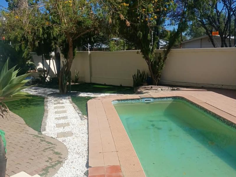 Outdoor pool area featuring a swimming pool with tiled coping, surrounded by a paved walkway, artificial grass, and a perimeter boundary wall.