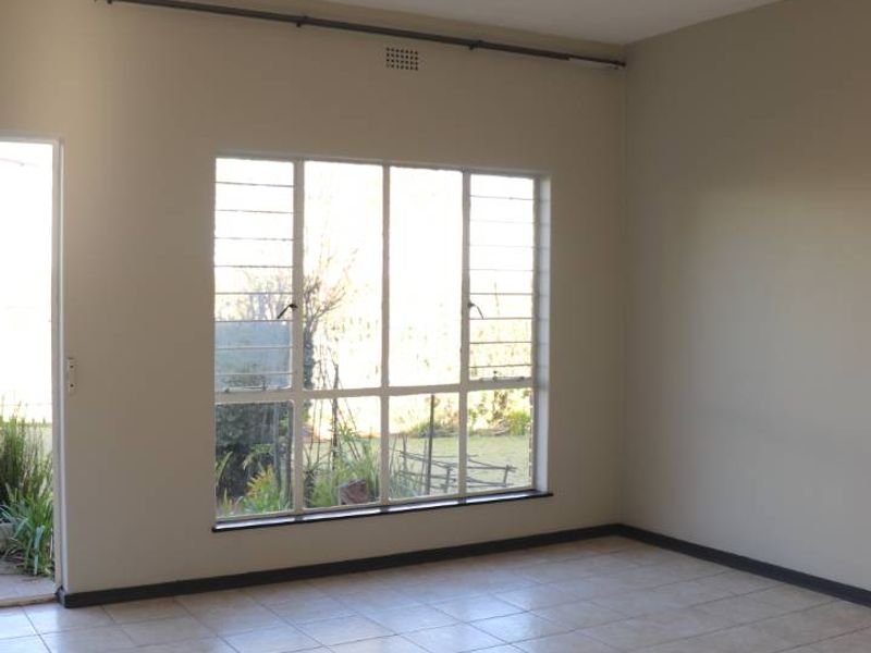 A bright, empty living room featuring neutral tiled flooring, a large steel-framed window, and a simple ceiling light fixture.