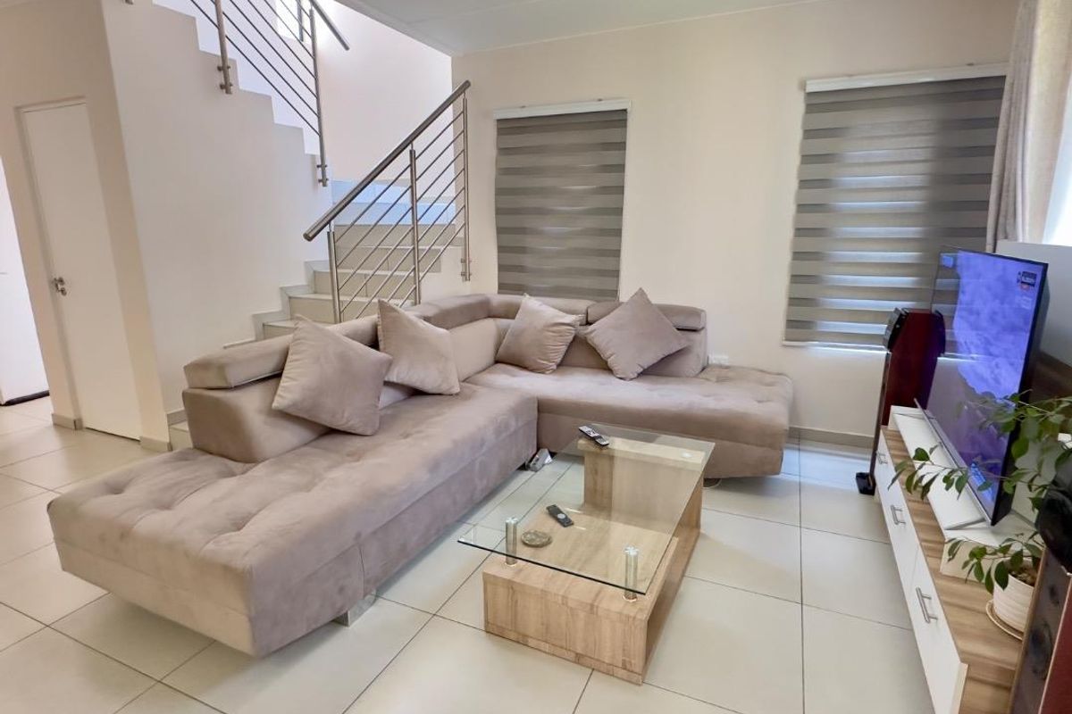 A bright and modern living room featuring tiled flooring, a staircase with a stainless steel balustrade, and dual roller blinds.