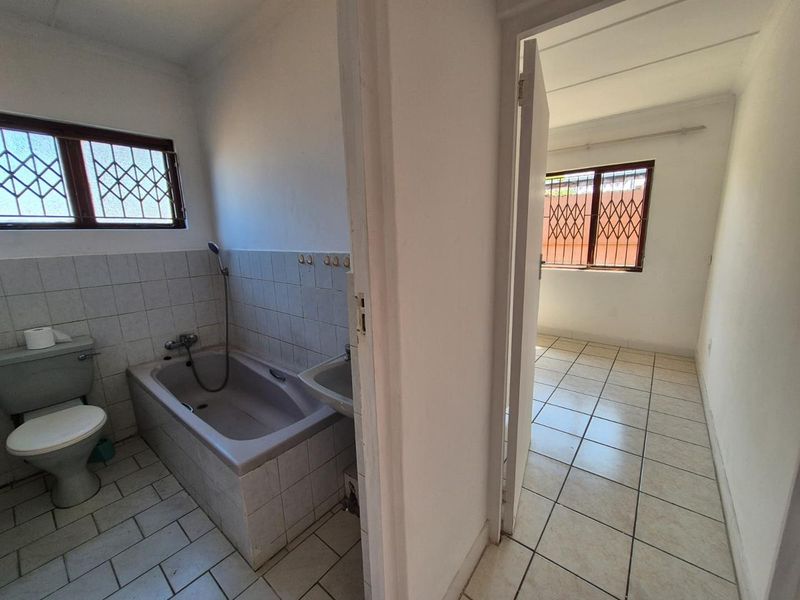 A dated bathroom featuring a bathtub, toilet, and washbasin, with tiled flooring and security bars on the window, adjacent to an empty room.