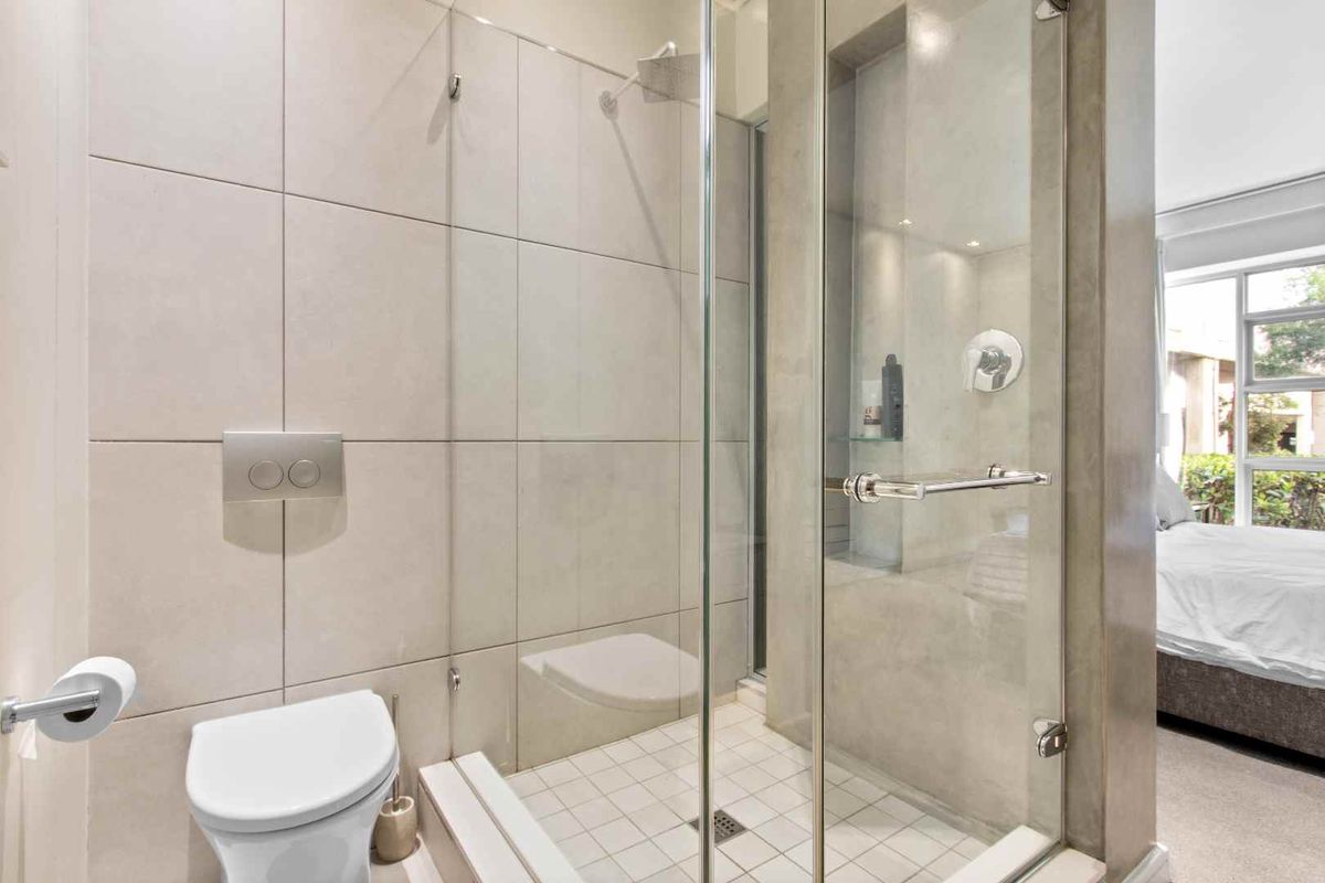 Modern bathroom featuring a glass-enclosed shower with a rainfall showerhead, wall-mounted toilet, and neutral tiled finishes, adjacent to a bedroom area.