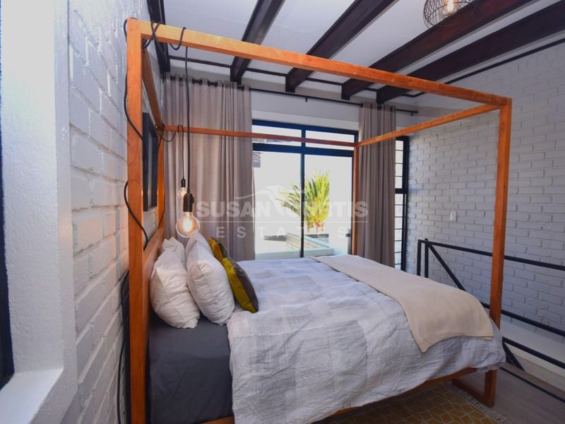 A well-lit bedroom featuring exposed timber ceiling beams, painted brick walls, and a sliding glass door providing access to an outdoor area.