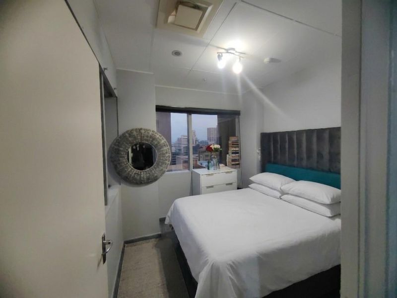A compact bedroom featuring a built-in headboard, ceiling lighting, and a window with an urban view.