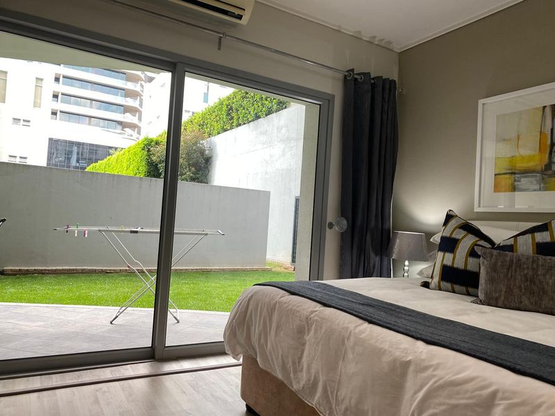 A bedroom featuring laminate flooring, a wall-mounted air conditioner, and large sliding glass doors providing direct access to a private patio and garden area.