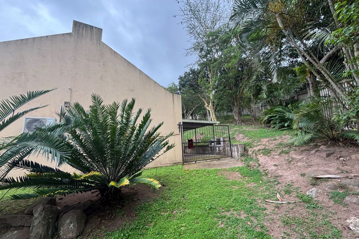Rear exterior view of the townhouse featuring a small concrete patio area, metal security gate, and sloped garden space with mature trees.