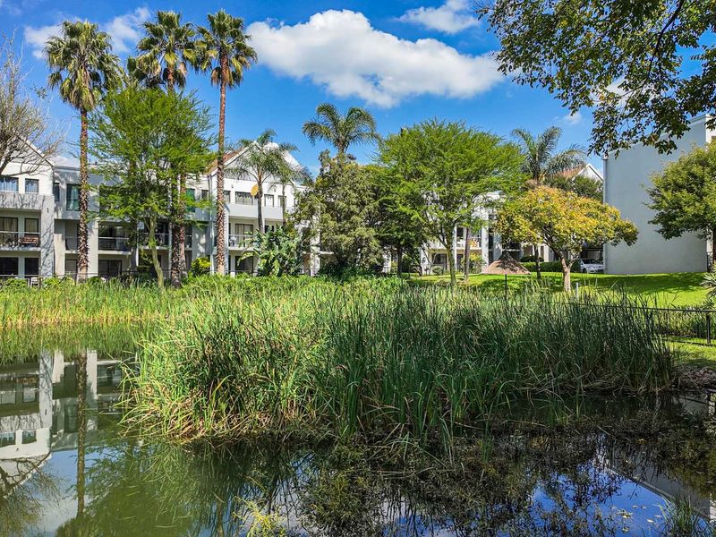 A scenic view of the apartment complex exterior featuring a tranquil pond, lush green gardens, and private balconies.