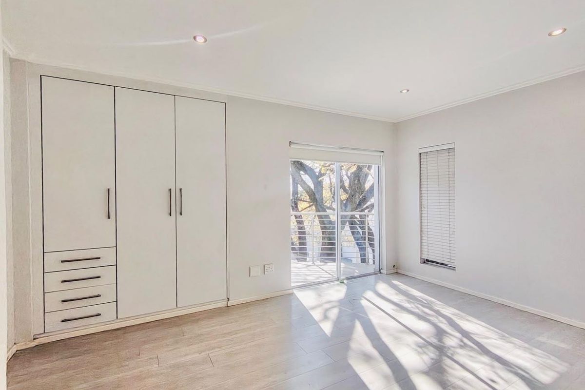 A bright, empty bedroom featuring modern laminate flooring, built-in cupboards, and a sliding door leading to an outdoor balcony area.