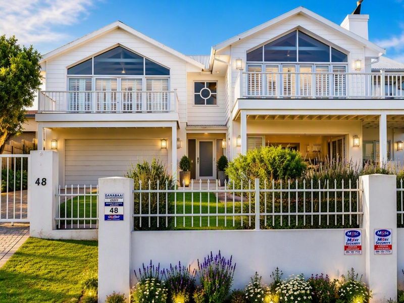 A beautifully maintained double-story home featuring a white facade, gabled roof, upper-level balconies, and a secure gated entrance with a paved driveway.