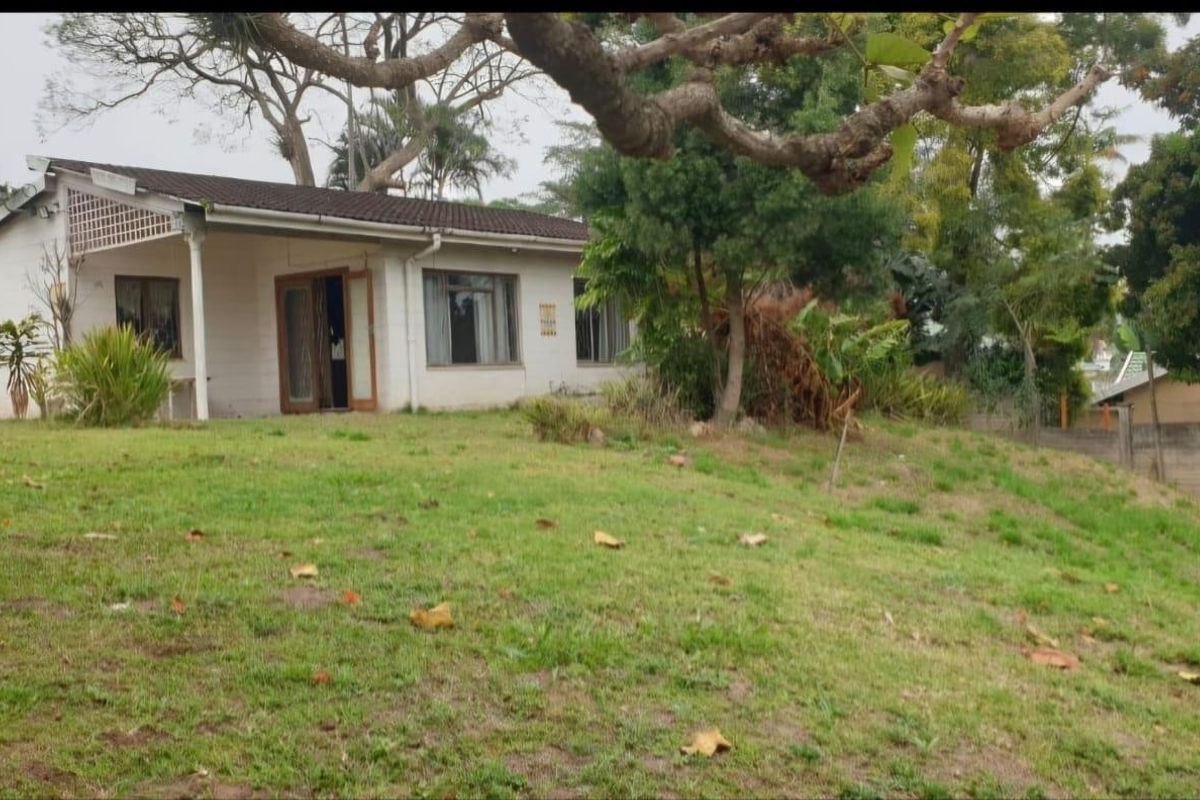 A rear view of a single-story house featuring a covered patio, sliding glass doors, and a large, sloped garden area with mature trees.