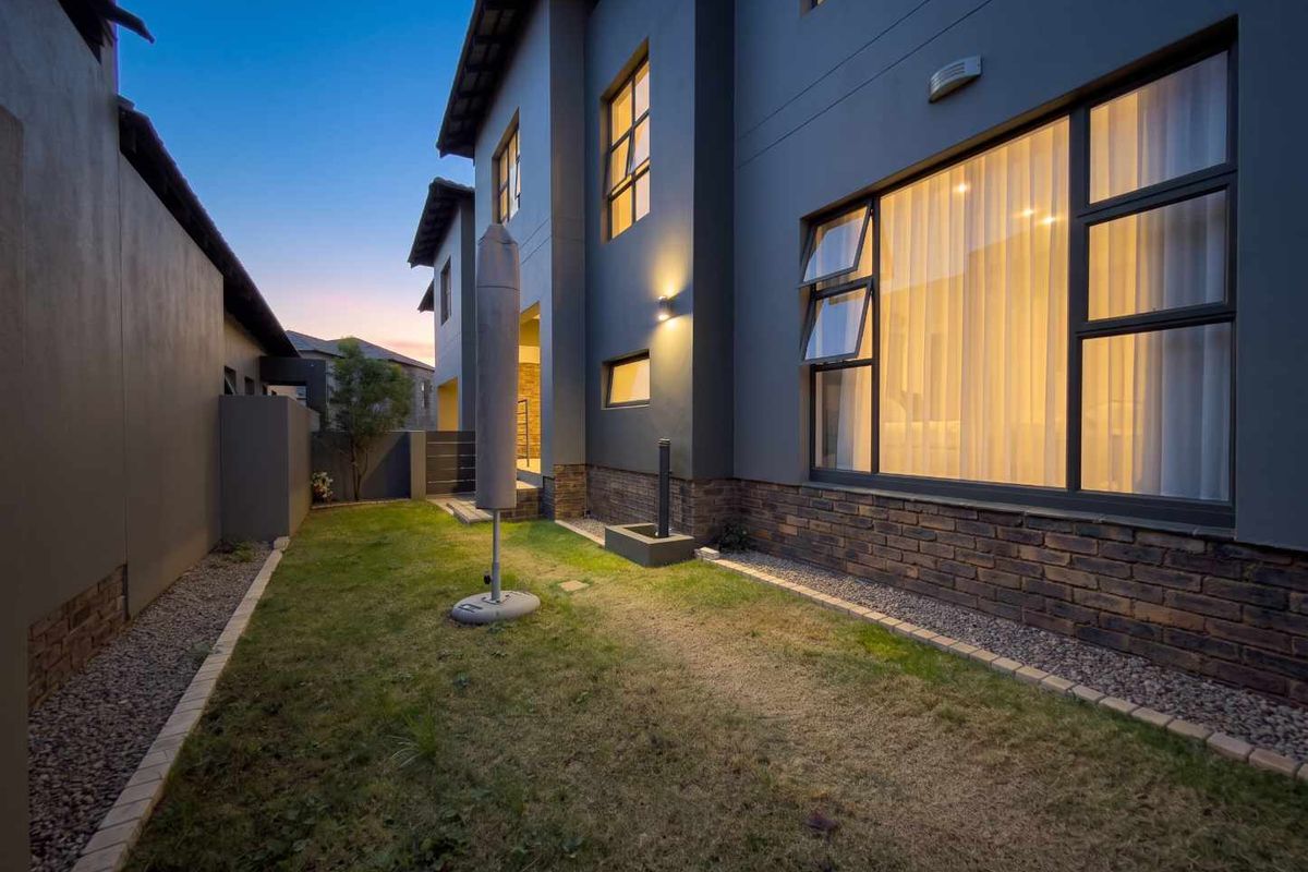 Rear exterior view of a modern townhouse featuring dark grey painted walls, brick accents, aluminium windows, and a landscaped garden area at dusk.