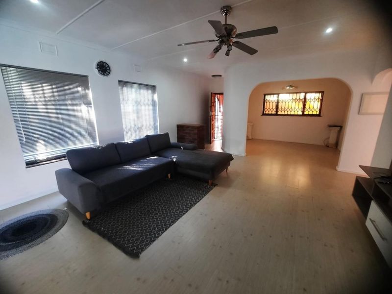 Spacious living room featuring laminate flooring, a ceiling fan, and large windows with venetian blinds, leading into an adjacent open-plan area.