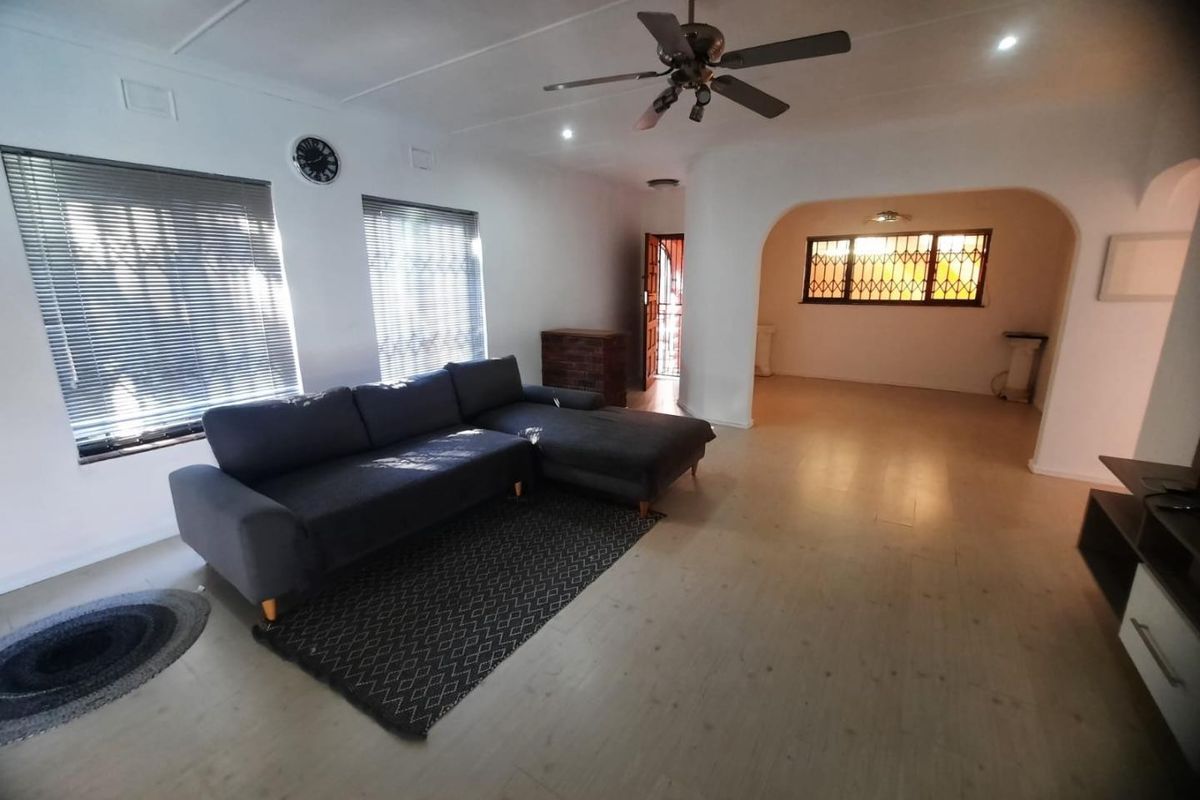 Spacious living room featuring laminate flooring, a ceiling fan, and large windows with venetian blinds, leading into an adjacent open-plan area.