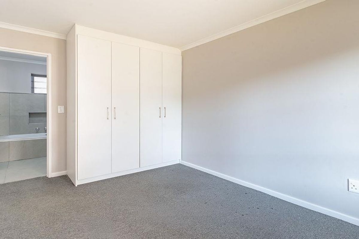 A clean, empty bedroom featuring neutral walls, carpeted flooring, and built-in cupboards, with an adjacent view into a tiled bathroom.
