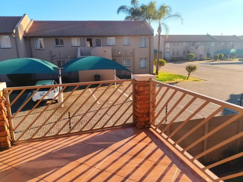 An elevated balcony with tiled flooring and metal balustrades, overlooking the complex parking area and paved driveway with shade cloth carports.