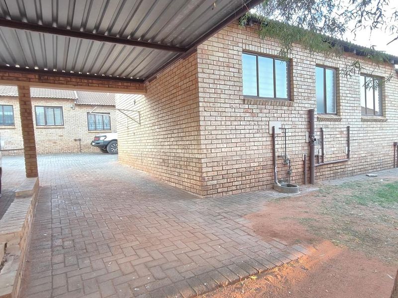 A covered carport area with brick construction and paved flooring, featuring exposed external plumbing and a corrugated metal roof structure.