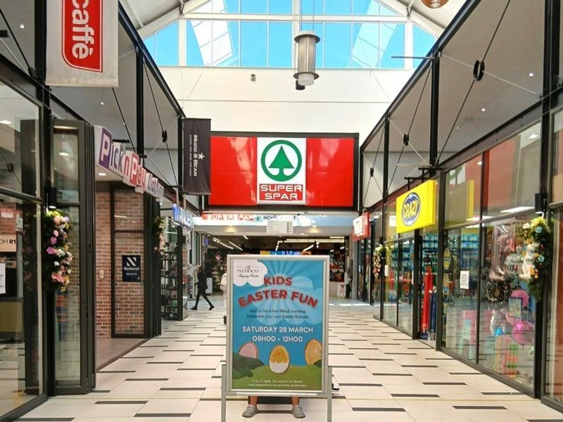 A bright, modern retail shopping centre corridor featuring high ceilings with skylights, glass-fronted retail units, and clean tiled flooring.