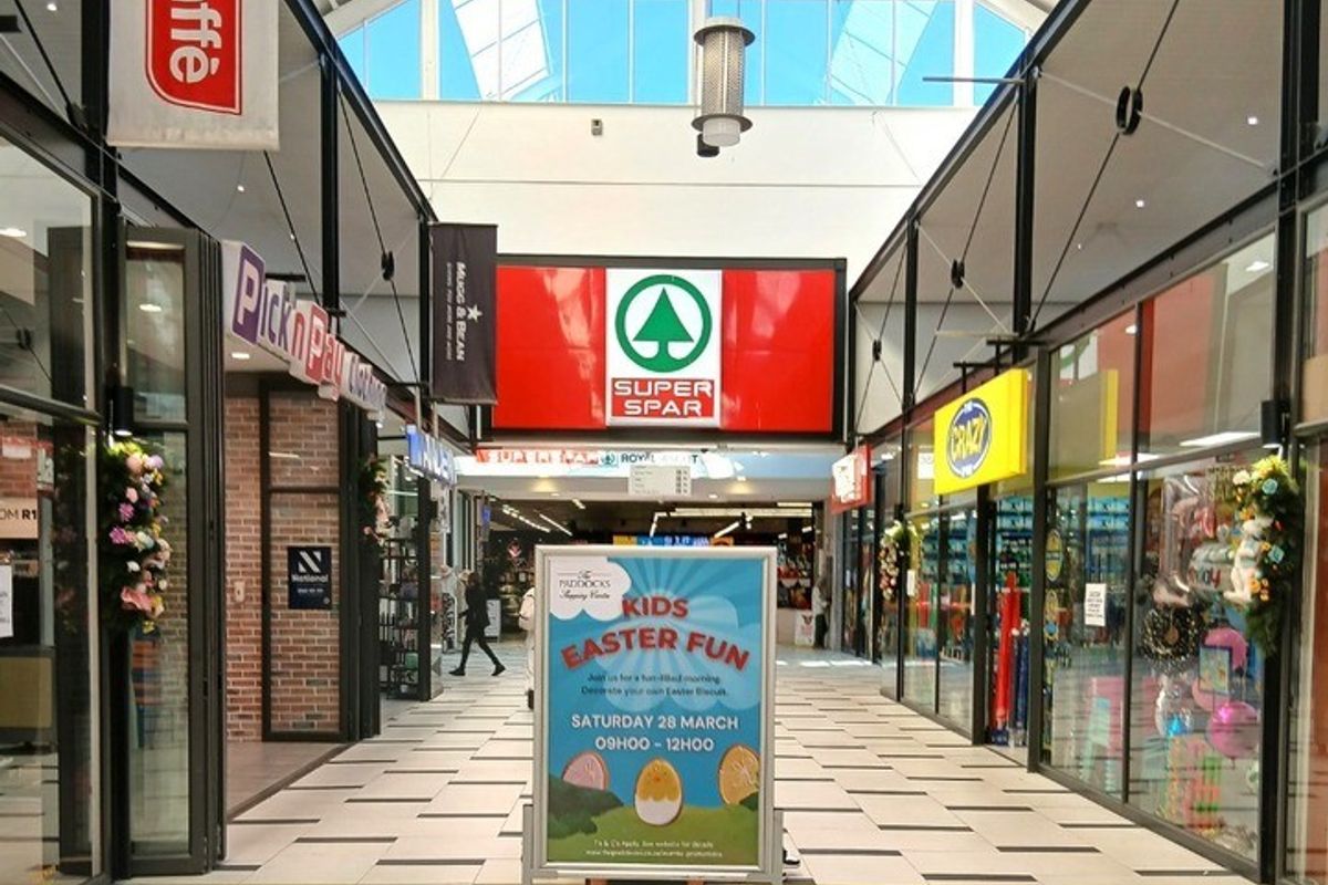 A bright, modern retail shopping centre corridor featuring high ceilings with skylights, glass-fronted retail units, and clean tiled flooring.