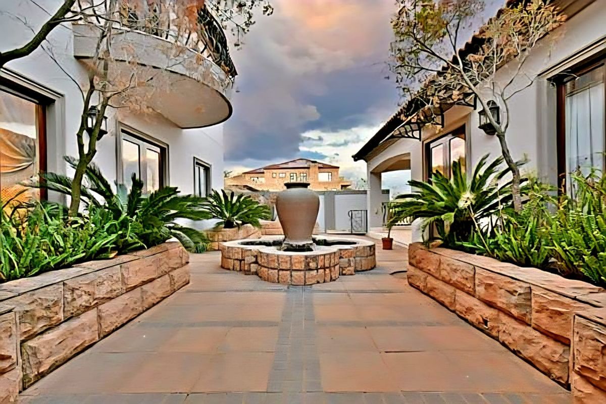A beautifully landscaped courtyard featuring a central stone fountain, paved walkways, and raised stone planters between two sections of the residence.