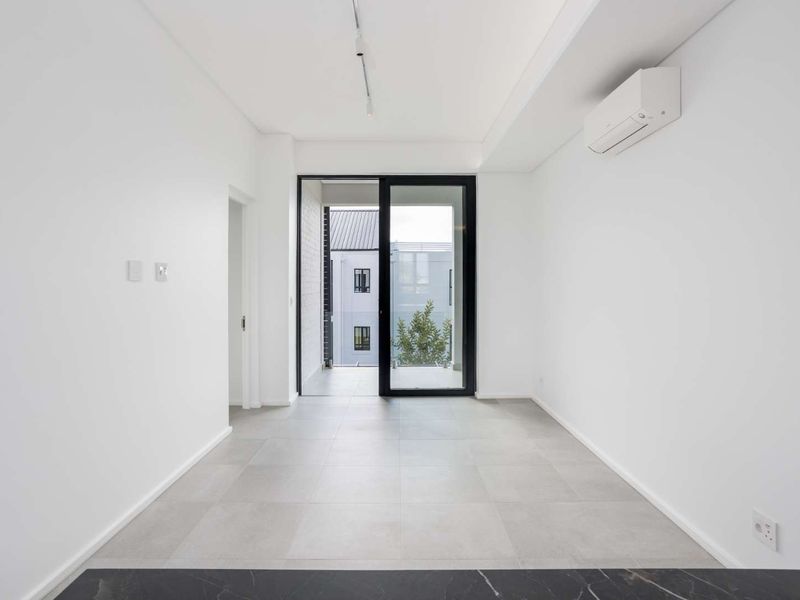 A bright, modern, and empty living area featuring neutral tiled flooring, a wall-mounted air conditioning unit, and a sliding glass door leading to a balcony.