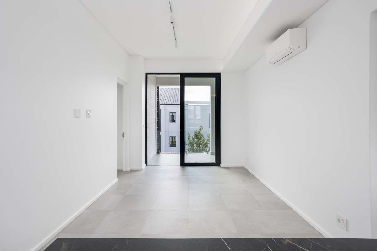 A bright, modern, and empty living area featuring neutral tiled flooring, a wall-mounted air conditioning unit, and a sliding glass door leading to a balcony.