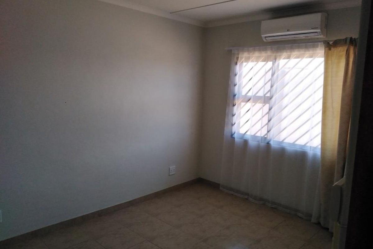 A neutral bedroom featuring tiled flooring, a wall-mounted air conditioning unit, and a window with security bars.